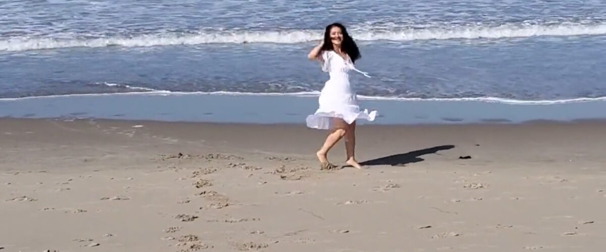 woman in a white dress with black hair on the beach with ocean waves behind her